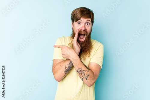 Young caucasian red-haired man isolated on blue background saying a gossip, pointing to side reporting something.