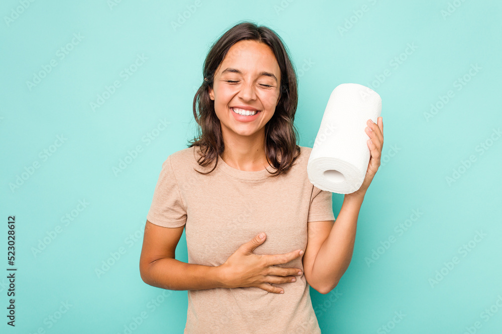 Young hispanic woman holding kitchen roll isolated on blue background laughing and having fun. Young hispanic woman holding kitchen roll isolated on blue background laughing and having fun.
