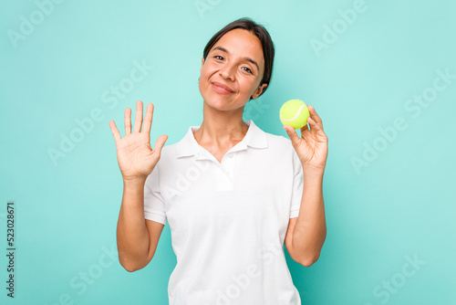 Young hispanic physiotherapy holding a tennis ball isolated on blue background smiling cheerful showing number five with fingers. © Asier