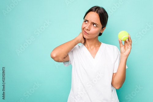 Young hispanic physiotherapy holding a tennis ball isolated on blue background touching back of head, thinking and making a choice. © Asier