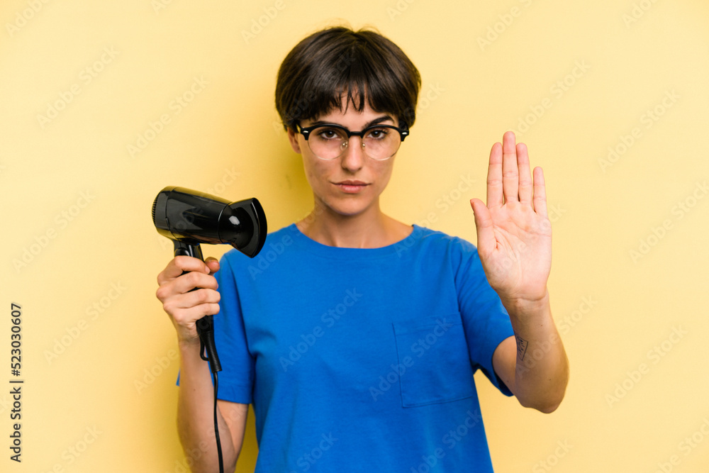 Young caucasian woman holding hairdryer isolated on yellow background standing with outstretched hand showing stop sign, preventing you. Young caucasian woman holding hairdryer isolated on yellow background standing with outstretched hand showing stop sign, preventing you.