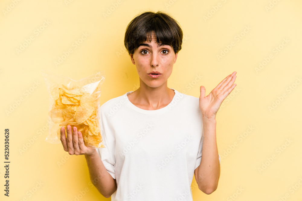 Young caucasian man holding crisps isolated on yellow background surprised and shocked. Young caucasian man holding crisps isolated on yellow background surprised and shocked.