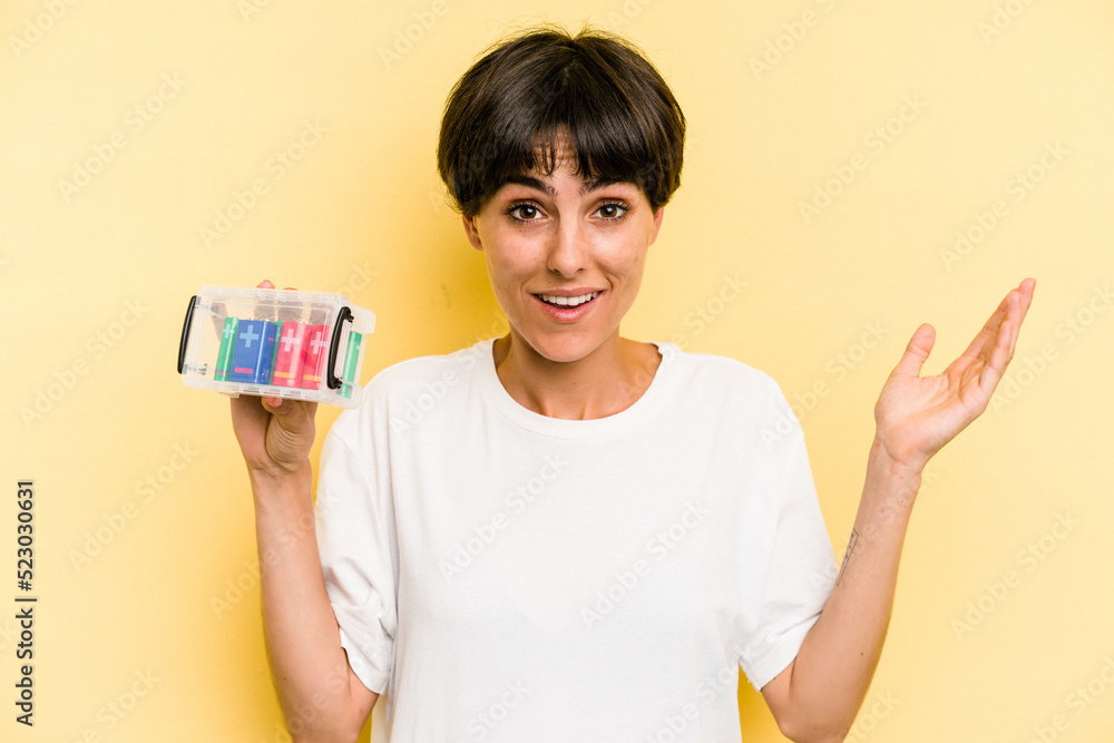 Young caucasian woman holding a batteries box isolated on yellow background receiving a pleasant surprise, excited and raising hands. Young caucasian woman holding a batteries box isolated on yellow background receiving a pleasant surprise, excited and raising hands.