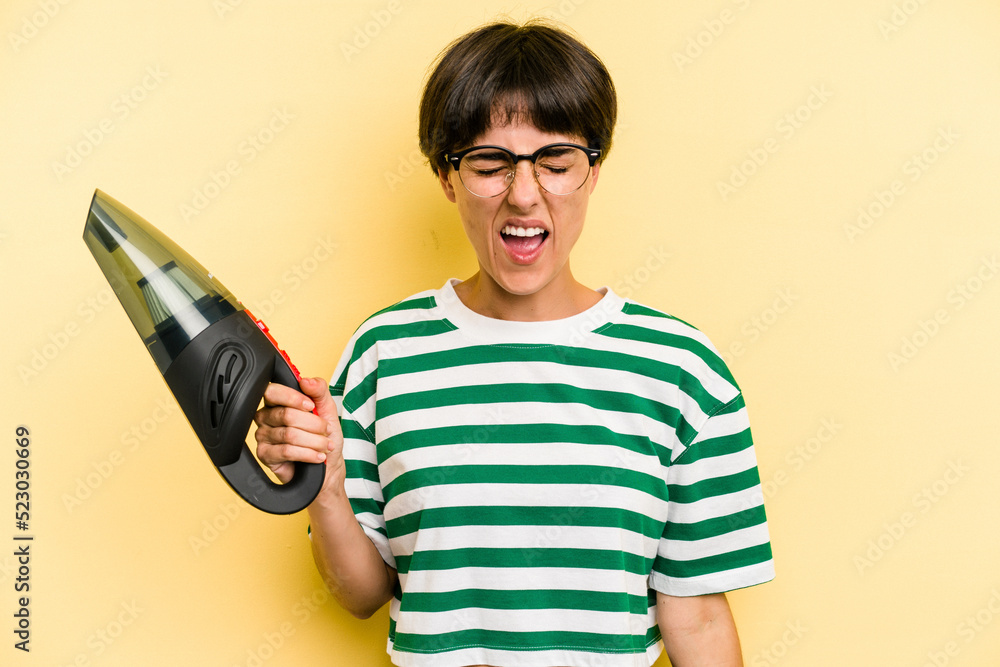 Young caucasian woman holding a hand vacuum cleaner isolated on yellow background screaming very angry and aggressive. Young caucasian woman holding a hand vacuum cleaner isolated on yellow background screaming very angry and aggressive.