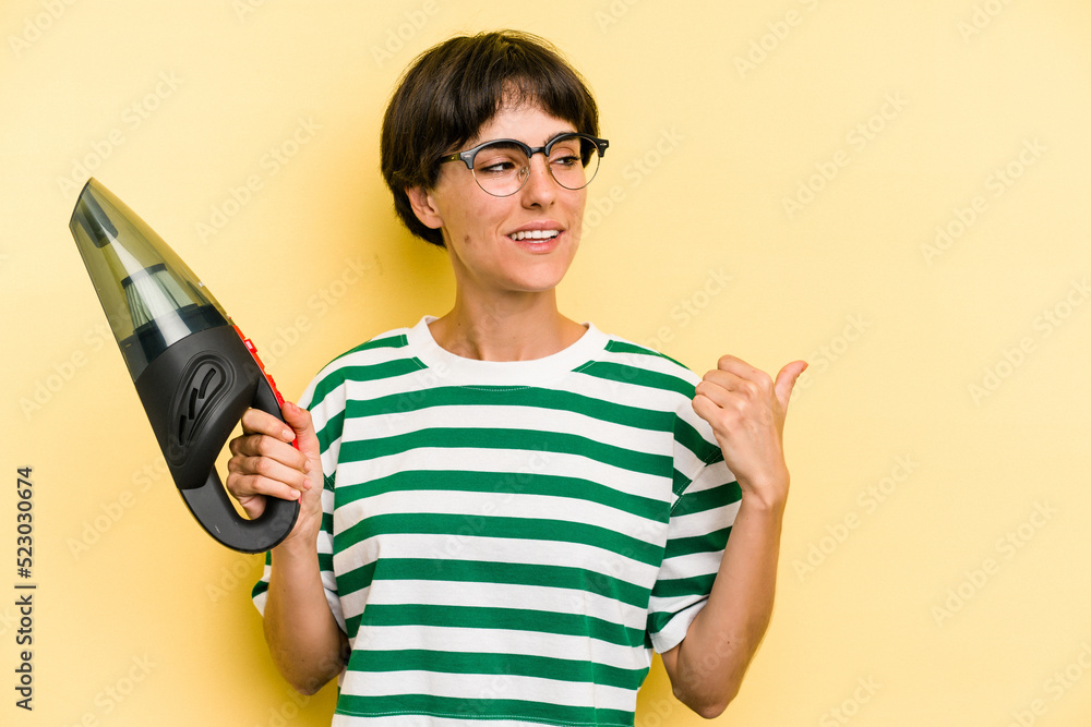 Young caucasian woman holding a hand vacuum cleaner isolated on yellow background points with thumb finger away, laughing and carefree. Young caucasian woman holding a hand vacuum cleaner isolated on yellow background points with thumb finger away, laughing and carefree.