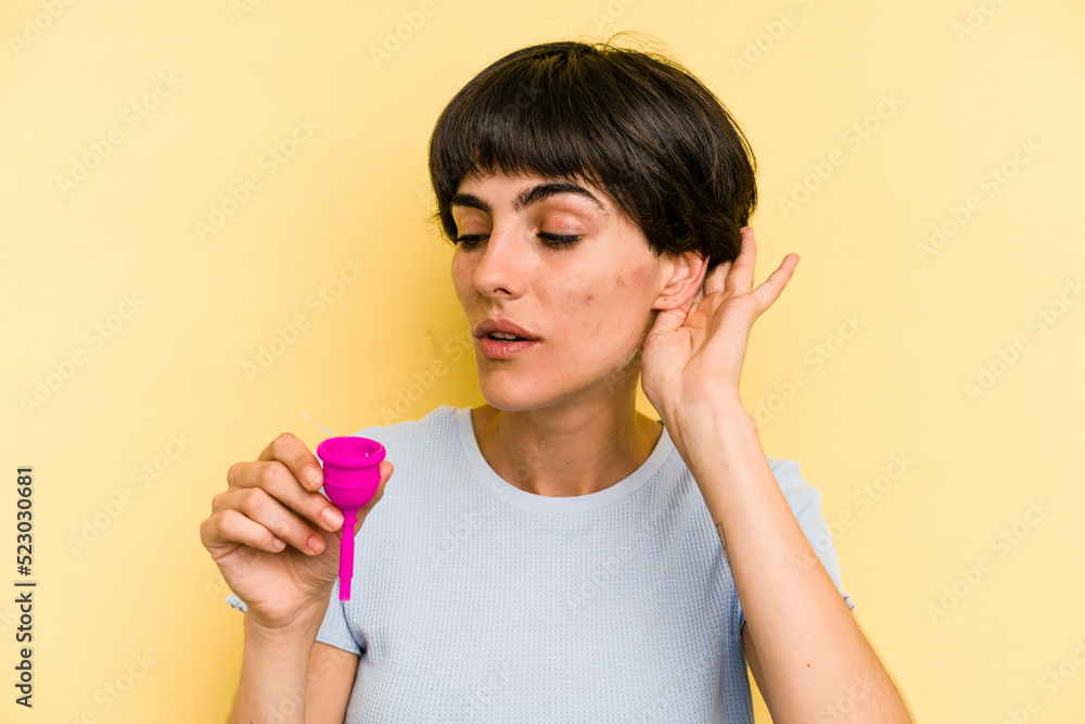 Young caucasian woman holding a menstrual cup isolated on yellow background trying to listening a gossip. Young caucasian woman holding a menstrual cup isolated on yellow background trying to listening a gossip.