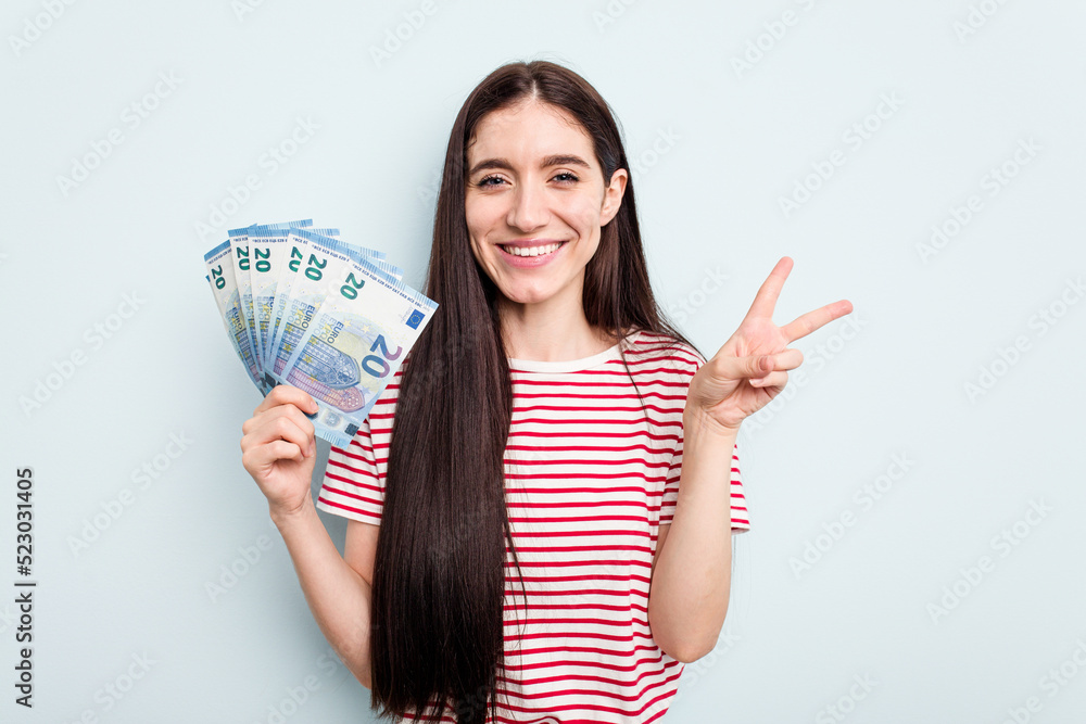 Young caucasian woman holding banknotes isolated on blue background joyful and carefree showing a peace symbol with fingers. Young caucasian woman holding banknotes isolated on blue background joyful and carefree showing a peace symbol with fingers.