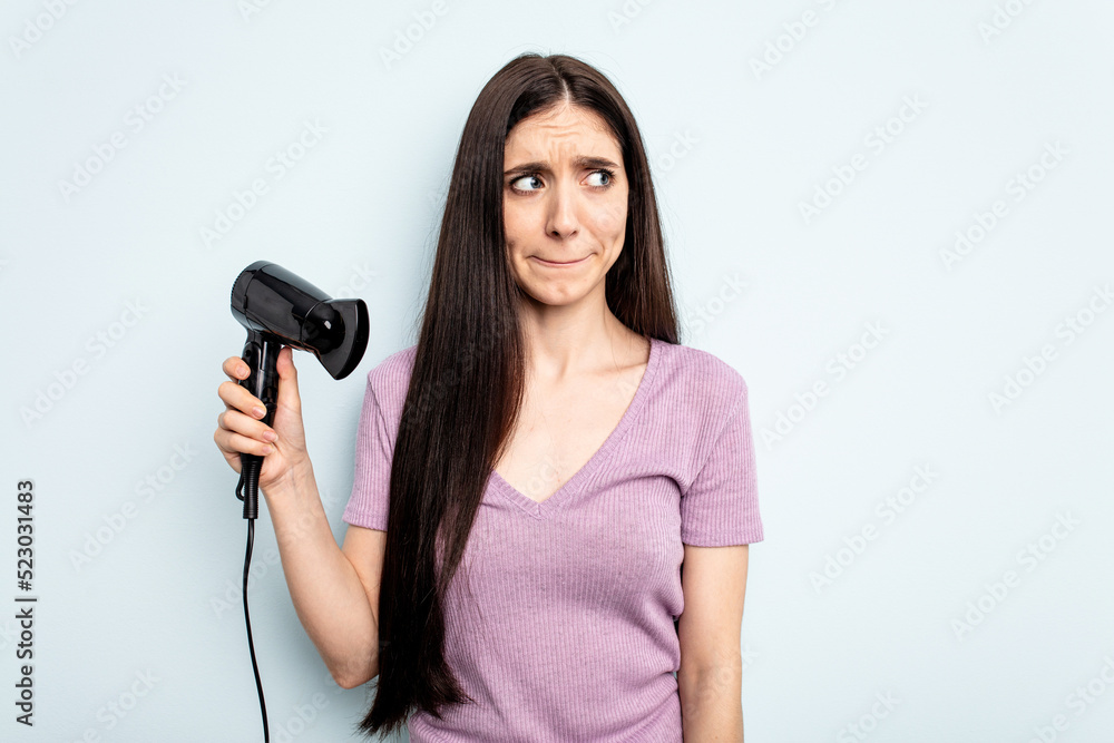 Young caucasian woman holding hairdryer isolated on blue background confused, feels doubtful and unsure. Young caucasian woman holding hairdryer isolated on blue background confused, feels doubtful and unsure.