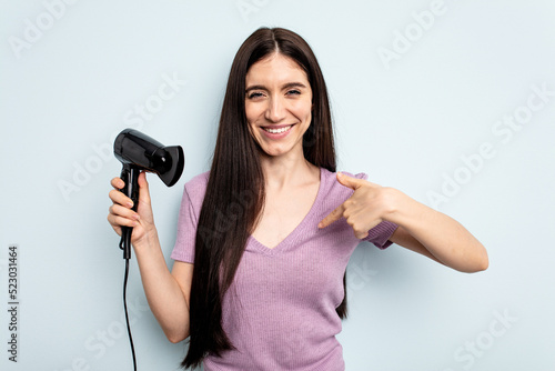 Young caucasian woman holding hairdryer isolated on blue background person pointing by hand to a shirt copy space, proud and confident