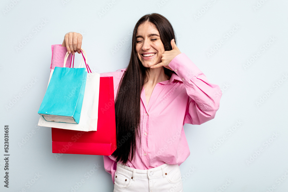 Young caucasian woman going to buy some clothes isolated on blue background showing a mobile phone call gesture with fingers. Young caucasian woman going to buy some clothes isolated on blue background showing a mobile phone call gesture with fingers.