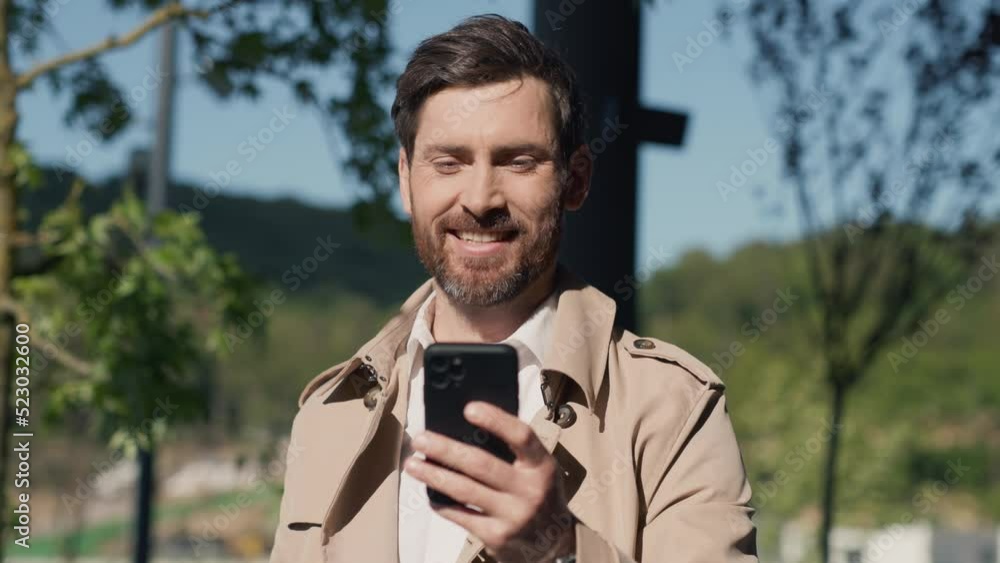 At sunlight happy smiling stylish handsome man with beard in coat uses smartphone stand in part. Close up. Slow motion At sunlight happy smiling stylish handsome man with beard in coat uses smartphone stand in part. Close up. Slow motion
