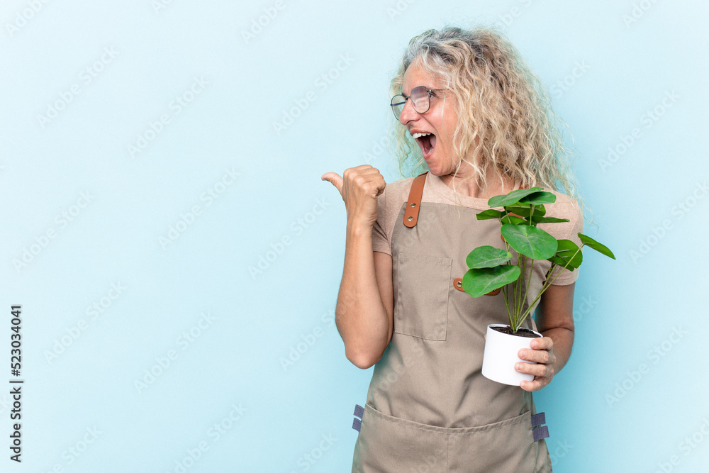 Middle age gardener woman holding a plant isolated on blue background points with thumb finger away, laughing and carefree. Middle age gardener woman holding a plant isolated on blue background points with thumb finger away, laughing and carefree.