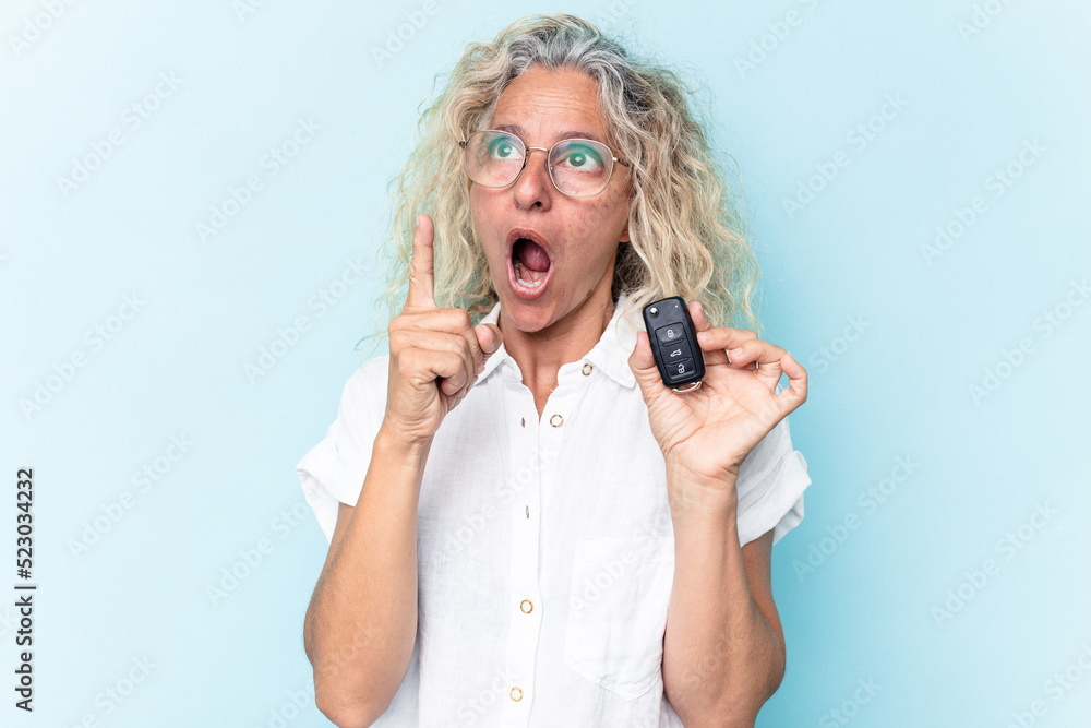 Middle age caucasian woman holding a car keys isolated on blue background pointing upside with opened mouth. Middle age caucasian woman holding a car keys isolated on blue background pointing upside with opened mouth.