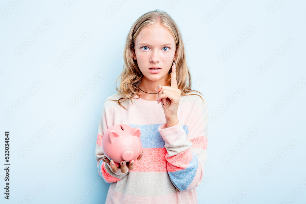 Caucasian teen girl holding a piggybank isolated on blue background having an idea, inspiration concept. Caucasian teen girl holding a piggybank isolated on blue background having an idea, inspiration concept.