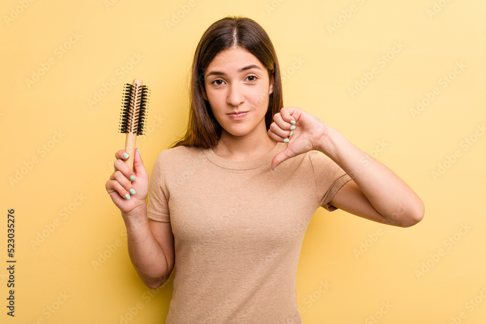 Young caucasian woman holding a brush hair isolated on yellow background showing a dislike gesture, thumbs down. Disagreement concept. Young caucasian woman holding a brush hair isolated on yellow background showing a dislike gesture, thumbs down. Disagreement concept.