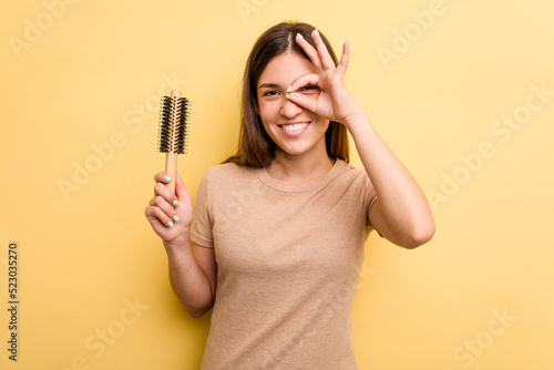 Young caucasian woman holding a brush hair isolated on yellow background excited keeping ok gesture on eye.