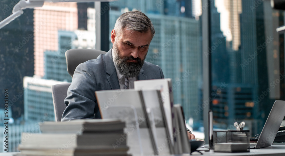 Portrait of 50s adult grey-haired Caucasian businessman signing documents in his office located in a skyscraper, business district buildings in the background Portrait of 50s adult grey-haired Caucasian businessman signing documents in his office located in a skyscraper, business district buildings in the background