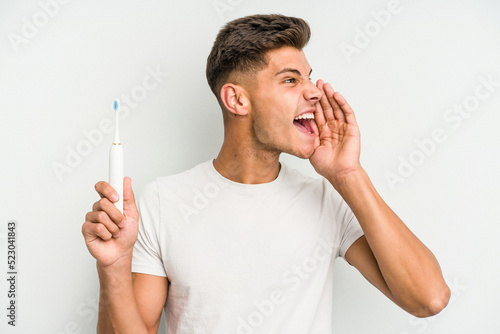 Young caucasian man holding a electric toothbrush isolated on white background shouting and holding palm near opened mouth.