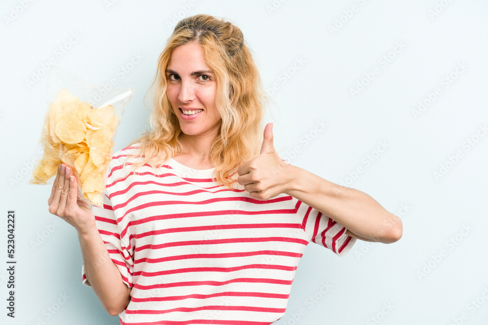 Young caucasian woman holding a bag of chips isolated on blue background smiling and raising thumb up Young caucasian woman holding a bag of chips isolated on blue background smiling and raising thumb up