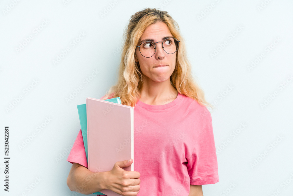 Young student caucasian woman holding books isolated on blue background confused, feels doubtful and unsure. Young student caucasian woman holding books isolated on blue background confused, feels doubtful and unsure.