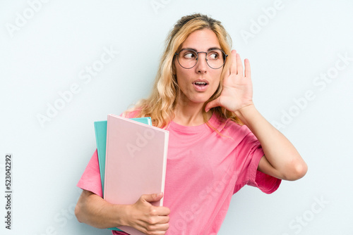 Young student caucasian woman holding books isolated on blue background trying to listening a gossip.