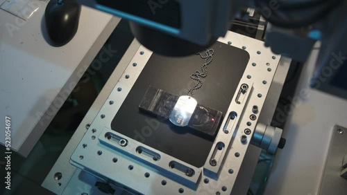 Laser engraving process of silver military soldier dog tag on chain of aluminum,top view. Laser technology workshop concept on gray metal surface,Ray movement from left to right,laser system details