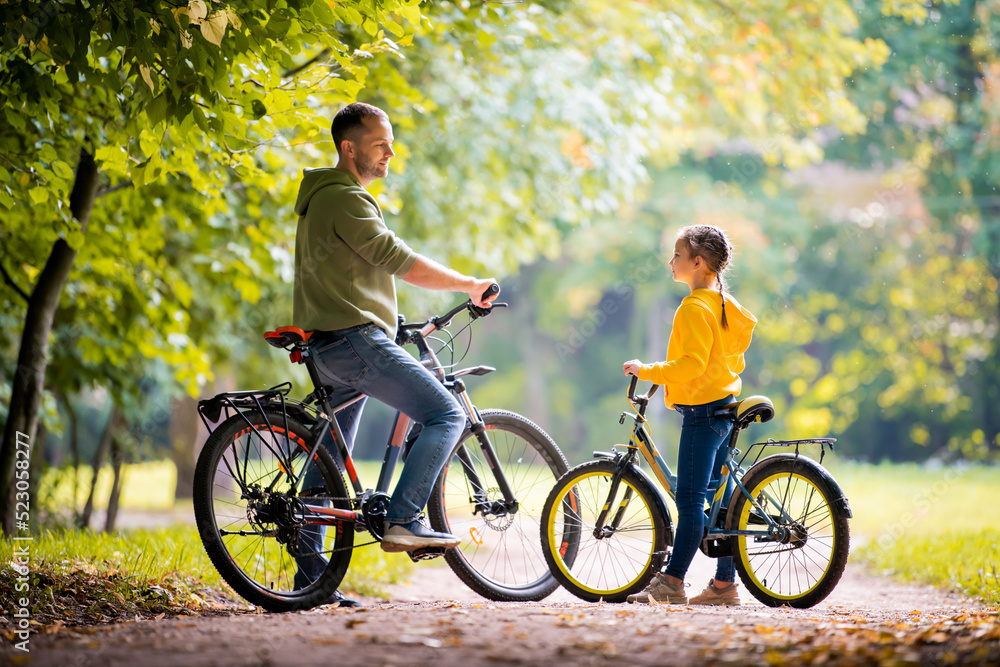Obraz premium Happy father and daughter walk with bicycles in the autumn park on a sunny day.