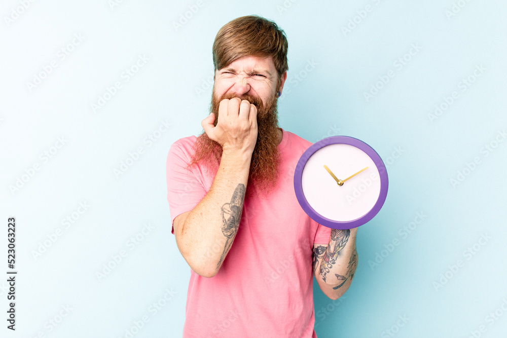 Young caucasian man holding a clock isolated on blue background biting fingernails, nervous and very anxious. Young caucasian man holding a clock isolated on blue background biting fingernails, nervous and very anxious.