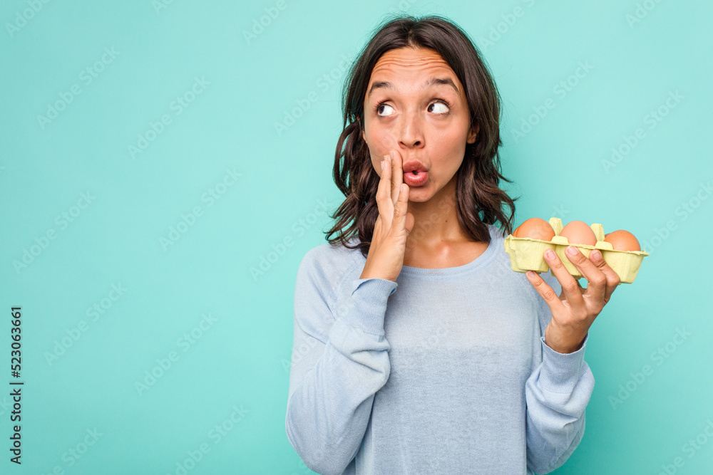 Young hispanic woman holding eggs isolated on blue background is saying a secret hot braking news and looking aside Young hispanic woman holding eggs isolated on blue background is saying a secret hot braking news and looking aside