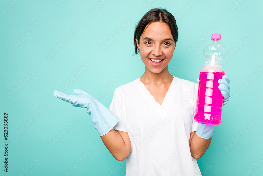 Young cleaner hispanic woman isolated on blue background showing a copy space on a palm and holding another hand on waist. Young cleaner hispanic woman isolated on blue background showing a copy space on a palm and holding another hand on waist.
