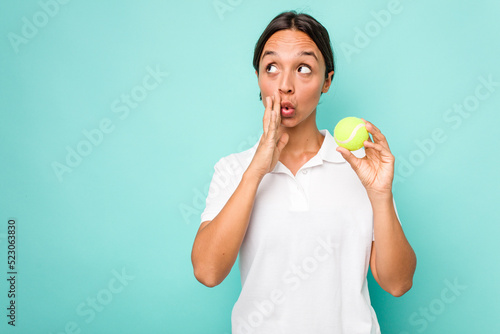 Young hispanic physiotherapy holding a tennis ball isolated on blue background is saying a secret hot braking news and looking aside © Asier