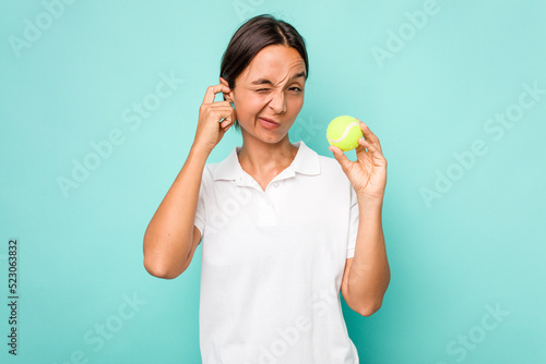 Young hispanic physiotherapy holding a tennis ball isolated on blue background covering ears with hands. © Asier