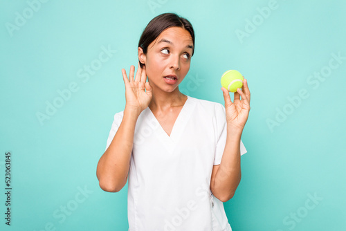 Young hispanic physiotherapy holding a tennis ball isolated on blue background trying to listening a gossip. © Asier