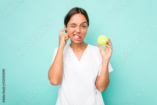 Young hispanic physiotherapy holding a tennis ball isolated on blue background covering ears with hands.