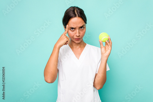 Young hispanic physiotherapy holding a tennis ball isolated on blue background pointing temple with finger, thinking, focused on a task. © Asier