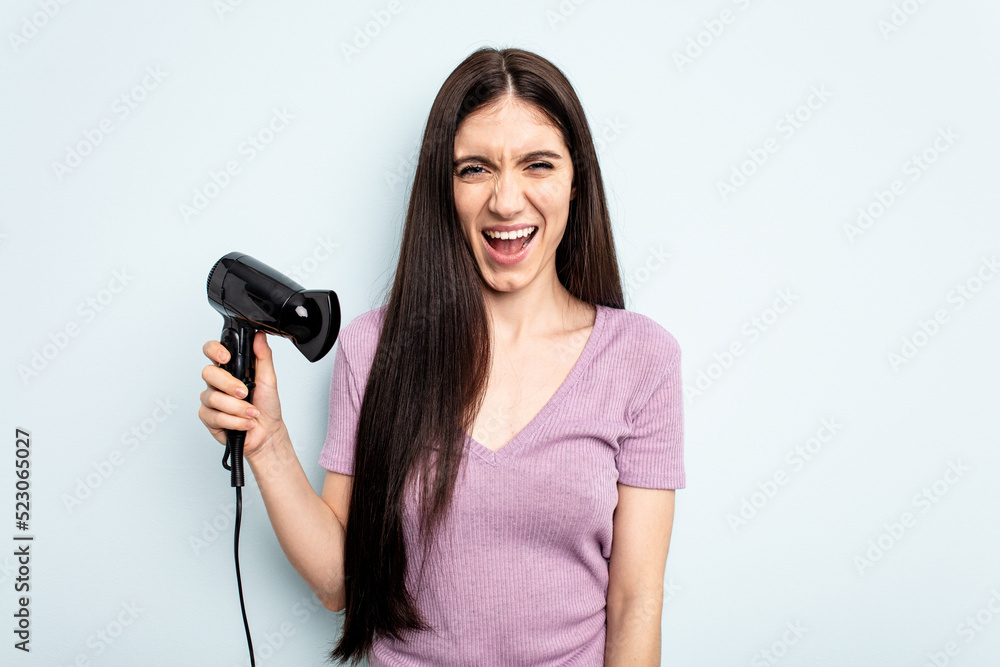Young caucasian woman holding hairdryer isolated on blue background screaming very angry and aggressive. Young caucasian woman holding hairdryer isolated on blue background screaming very angry and aggressive.