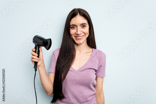 Young caucasian woman holding hairdryer isolated on blue background happy, smiling and cheerful.