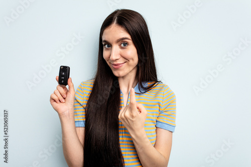 Young caucasian woman holding car keys isolated on blue background pointing with finger at you as if inviting come closer.
