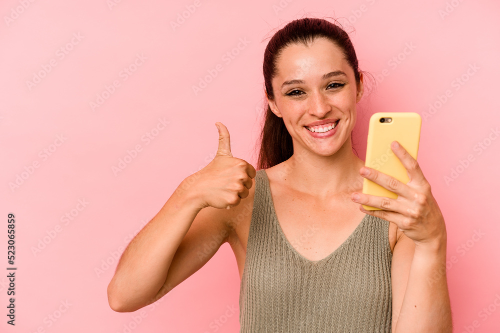 Young caucasian woman holding mobile phone isolated on pink background smiling and raising thumb up Young caucasian woman holding mobile phone isolated on pink background smiling and raising thumb up