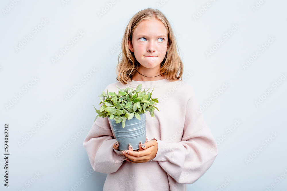 Caucasian teen girl holding a plant isolated on blue background confused, feels doubtful and unsure. Caucasian teen girl holding a plant isolated on blue background confused, feels doubtful and unsure.