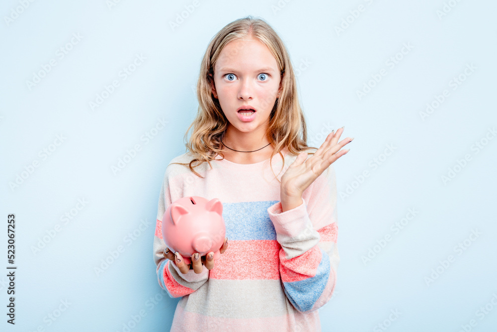 Caucasian teen girl holding a piggybank isolated on blue background surprised and shocked. Caucasian teen girl holding a piggybank isolated on blue background surprised and shocked.