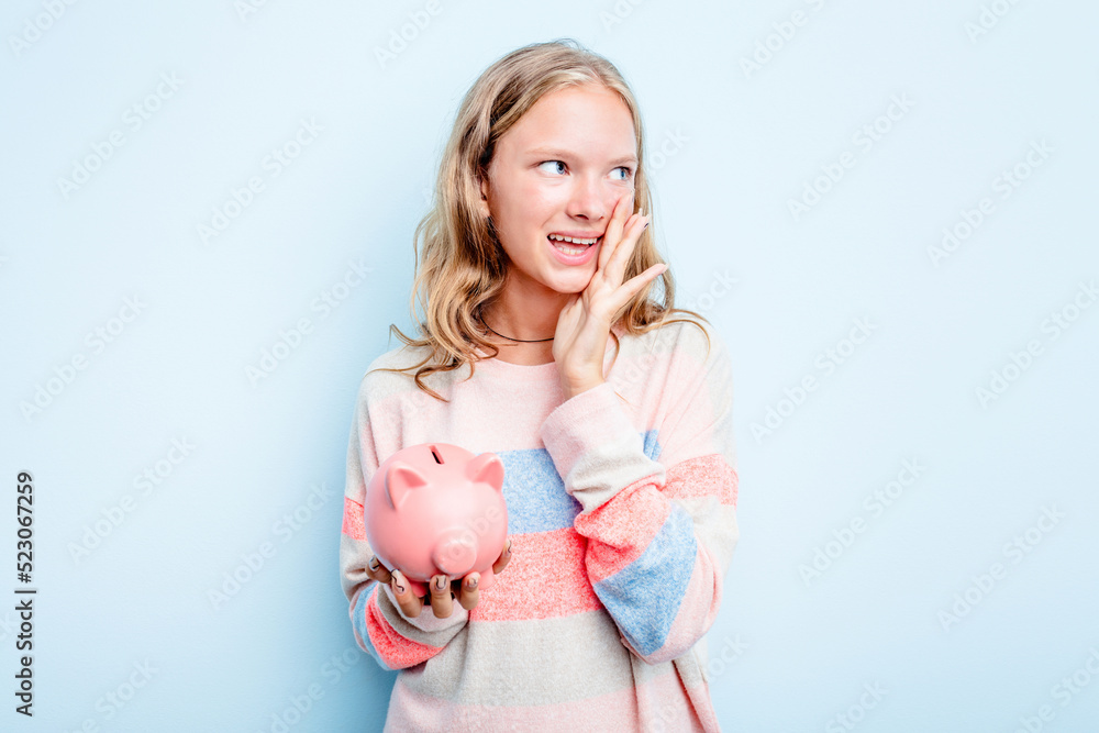 Caucasian teen girl holding a piggybank isolated on blue background shouting and holding palm near opened mouth. Caucasian teen girl holding a piggybank isolated on blue background shouting and holding palm near opened mouth.