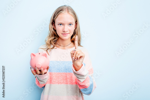 Caucasian teen girl holding a piggybank isolated on blue background showing number one with finger.