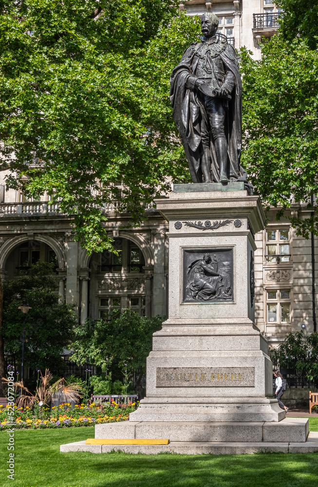 London, UK - July 4, 2022: Whitehall Gardens.  Black bromze Sir Henry Bartle Edward Frere statue on stone pedestal with Pro Patria mural backed by green foliage. Lawn and yellow flowers.