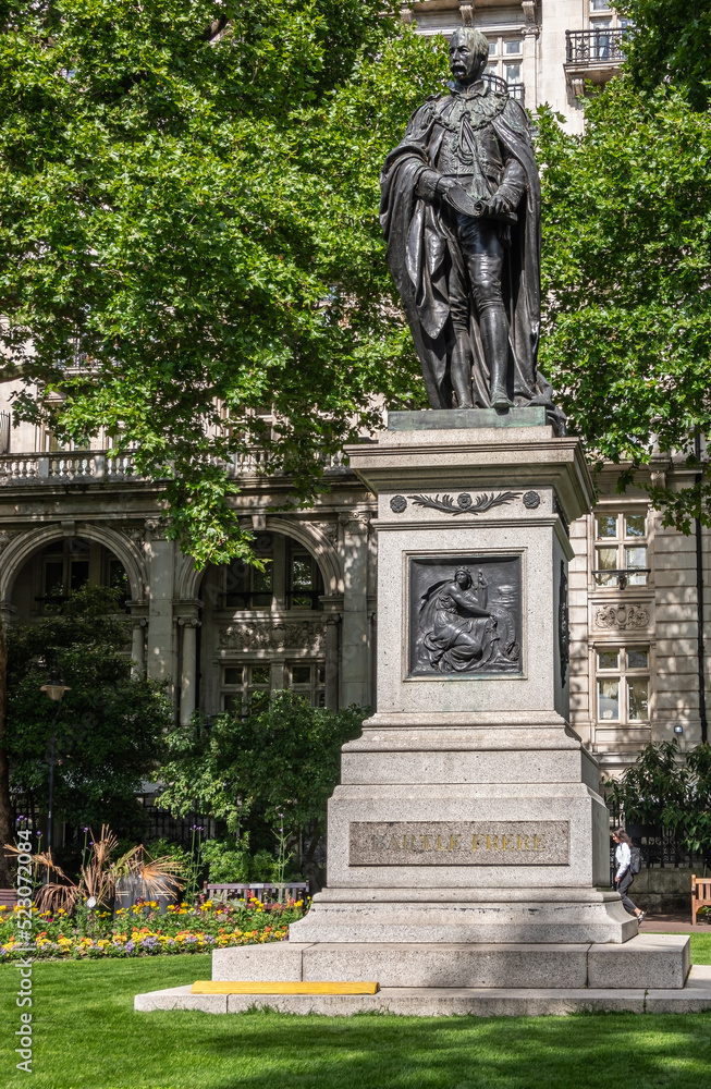 Obraz premium London, UK - July 4, 2022: Whitehall Gardens. Black bromze Sir Henry Bartle Edward Frere statue on stone pedestal with Pro Patria mural backed by green foliage. Lawn and yellow flowers.