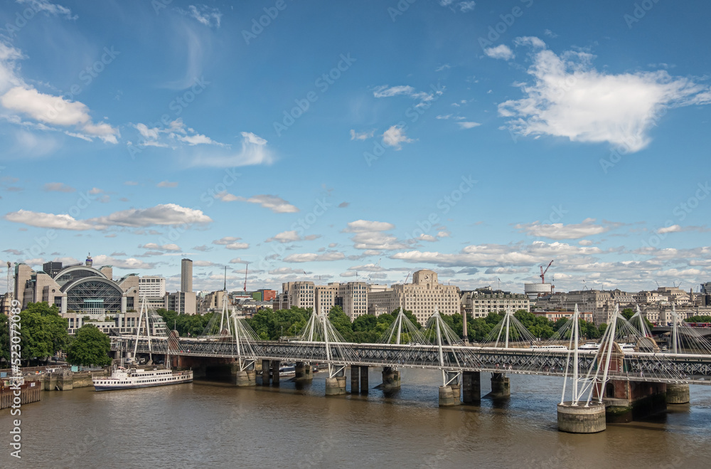 Fototapeta premium London, UK - July 4, 2022: Golden Jubilee bridges on both sides of Hungerford Bridge spanning brown water Thame river under blue cloudscape. Charing Cross railway station and cityscape.