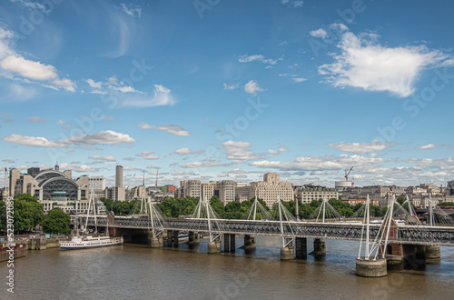 Платно London, UK - July 4, 2022: Golden Jubilee bridges on both sides of Hungerford Bridge spanning brown water Thame river under blue cloudscape