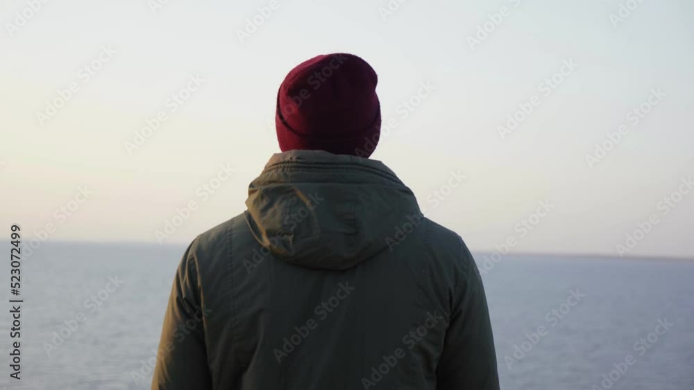 Hiking hobby. Back view of alone man backpacker in red hat looks sea Hiking hobby. Back view of alone man backpacker in red hat looks sea