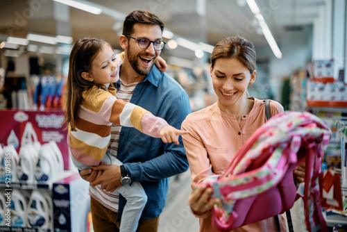 Fotografie Happy little girl points at backpack while buying school supplies with her parents in supermarket