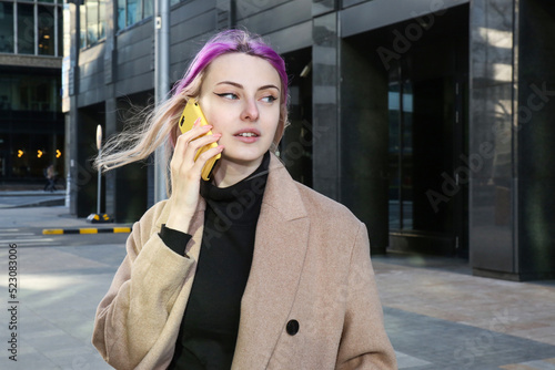 blonde with lilac strands of hair in a beige coat and glasses on the street with a phone in her hand